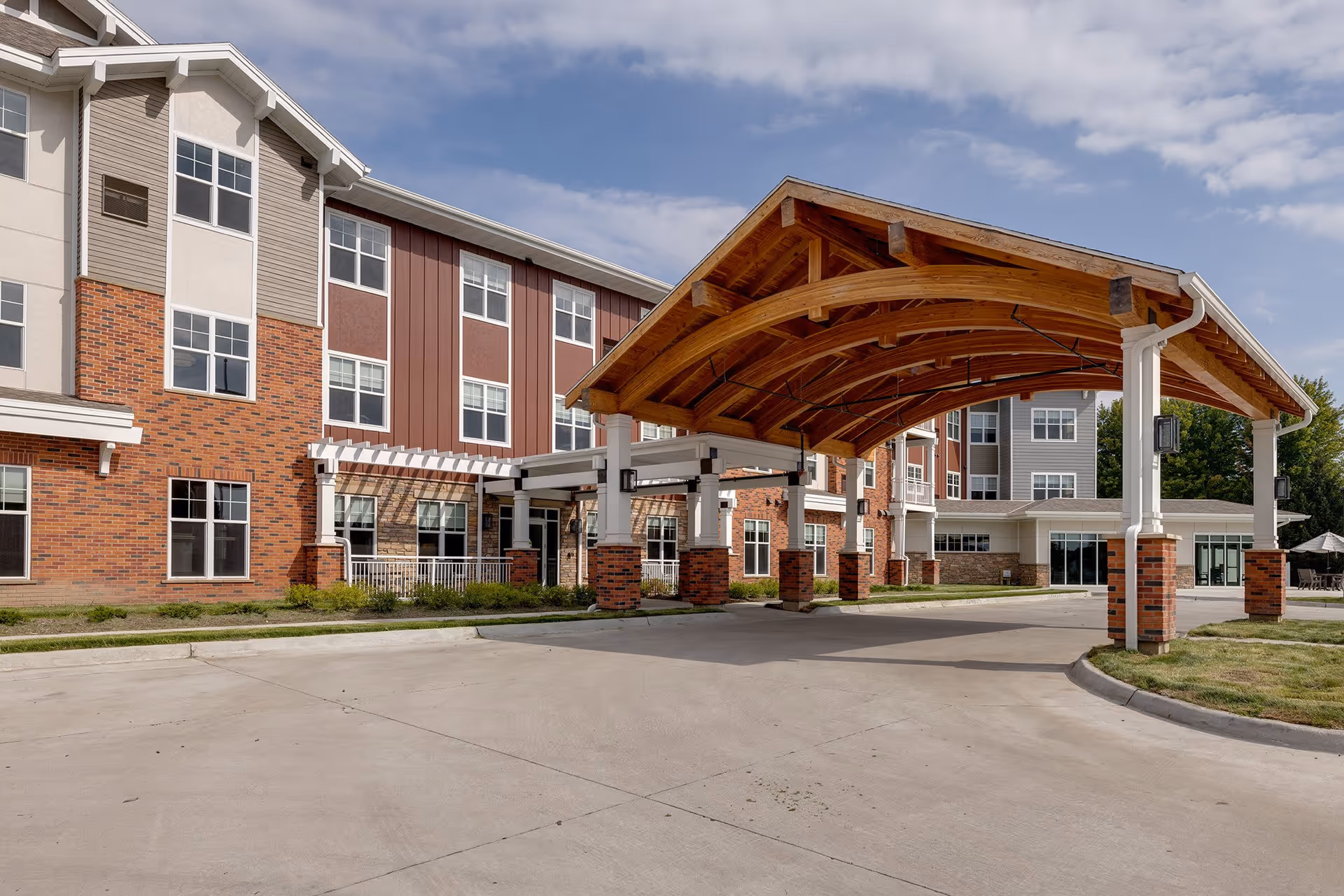 Exterior view of Meadowview of Johnston senior living facility showing a large covered entrance with wooden beams and brick pillars, a multi-story building with a mix of brick and siding, and a clear sky with some clouds.