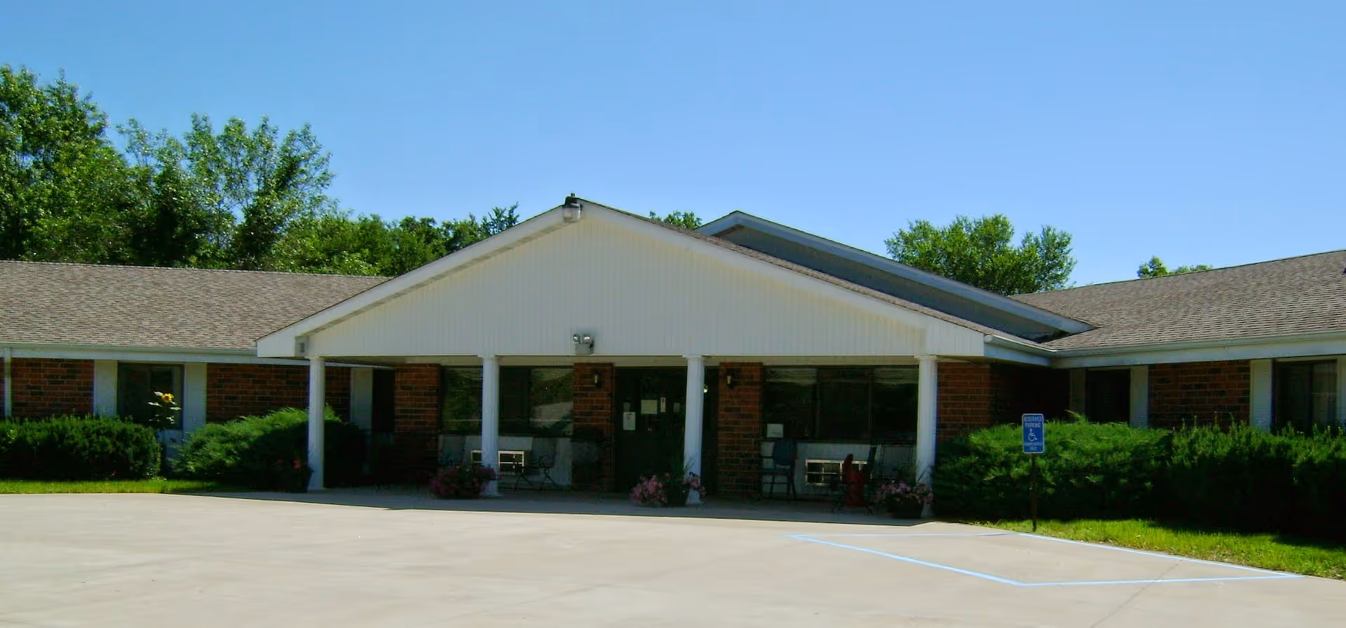Exterior view of a single-story brick building with a covered entrance supported by white columns. There are bushes and greenery around the building and a clear blue sky above. A handicapped parking sign is visible near the entrance.