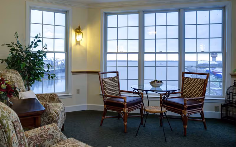 A cozy sitting area with two wooden chairs and a small round glass table between them, positioned in front of large windows overlooking a body of water. There are two upholstered armchairs with floral patterns and a wooden side table with a plant and flowers on it. The room has soft lighting from a wall-mounted lamp.