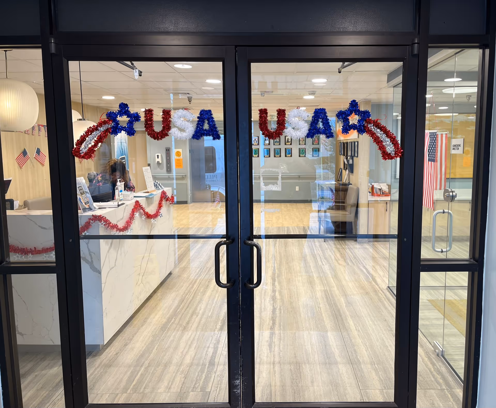 Glass double doors decorated with red, white, and blue USA-themed garlands. Inside, a reception desk with a person sitting behind it is visible, along with American flags on the walls and a waiting area with chairs. The floor is light-colored wood or laminate, and the area is well-lit with ceiling lights.