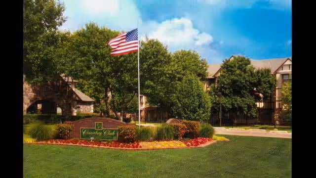 Exterior view of the Country Club At Woodland Hills facility showing a landscaped area with green grass, colorful flower beds, an American flag on a flagpole, and trees in front of a multi-story building under a blue sky with some clouds.