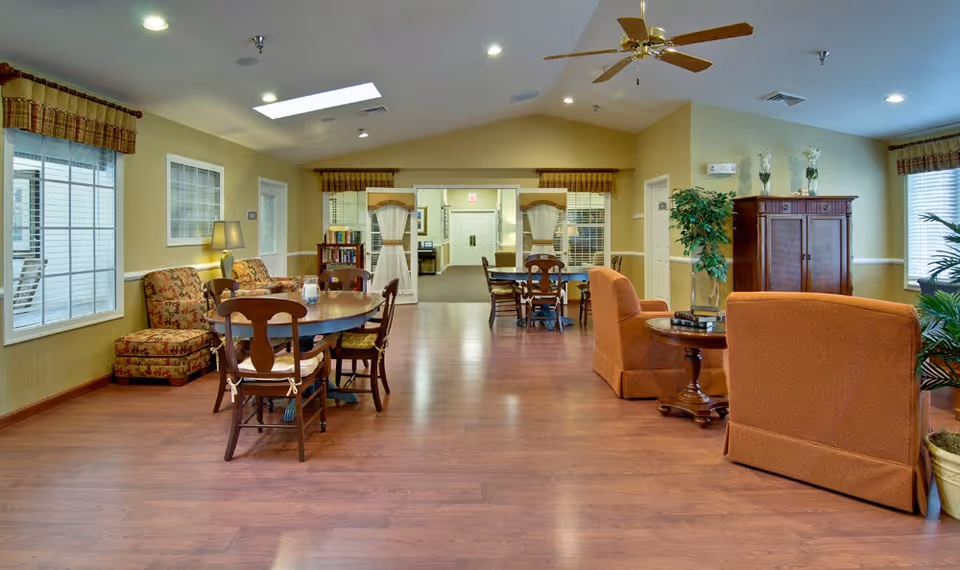 A spacious and well-lit common area in a senior living facility with wooden flooring, multiple seating arrangements including cushioned armchairs, floral-patterned chairs, and wooden dining tables with chairs. The room has large windows with valances, a ceiling fan, potted plants, and a wooden cabinet. The walls are painted a warm beige color, and there is a skylight on the ceiling.