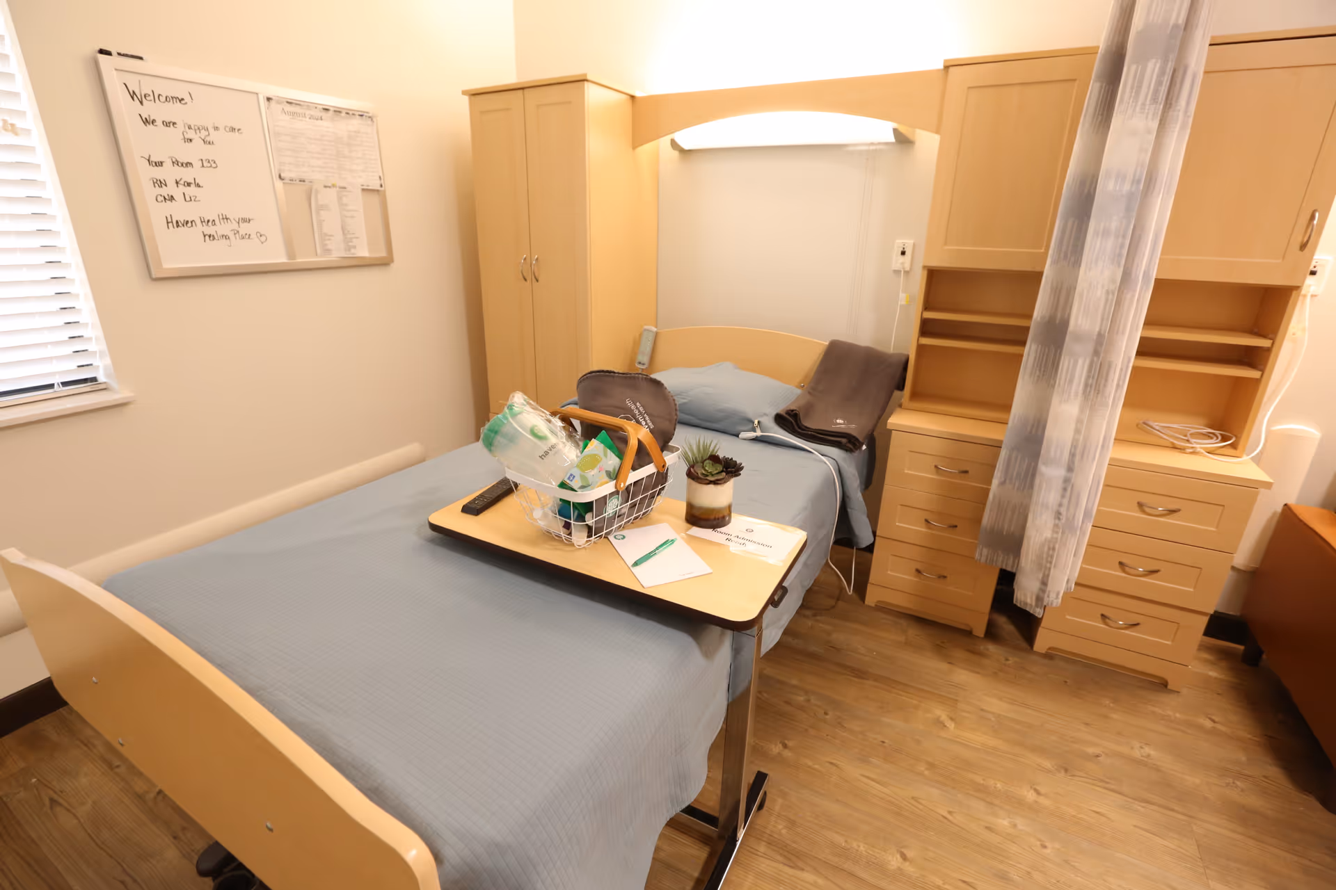 A small, neatly arranged bedroom in a senior living facility with a single bed covered in a light blue blanket. A wooden overbed table holds a basket with various items, a remote control, a small plant, and some papers. The room has wooden furniture including a wardrobe, a chest of drawers, and a shelving unit. A whiteboard with handwritten notes is mounted on the wall near a window with blinds. The floor is wooden, and a privacy curtain is partially visible.