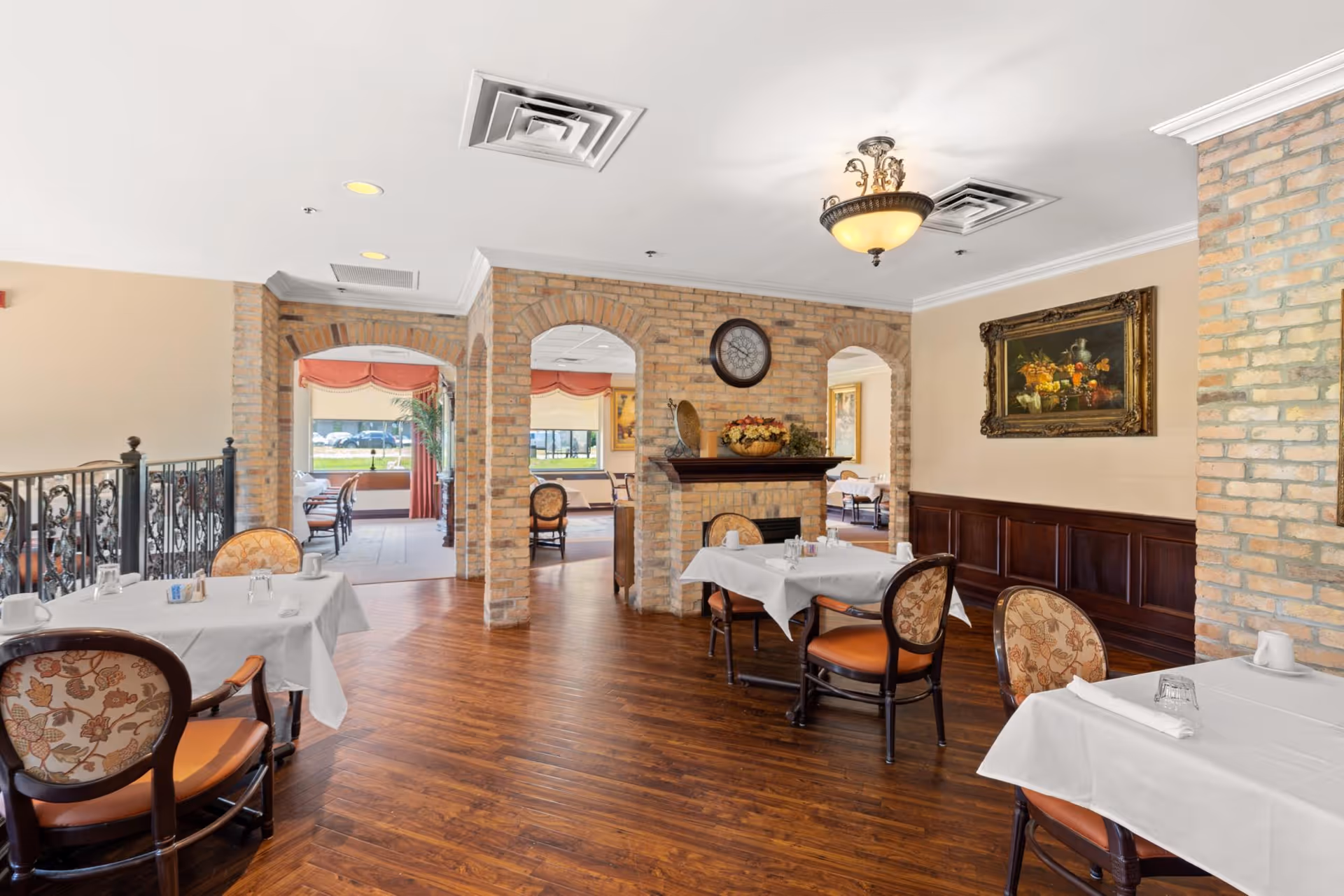 Elegant dining room with white-clothed tables and upholstered chairs, brick archways, a decorative fireplace with a clock, and framed artwork.