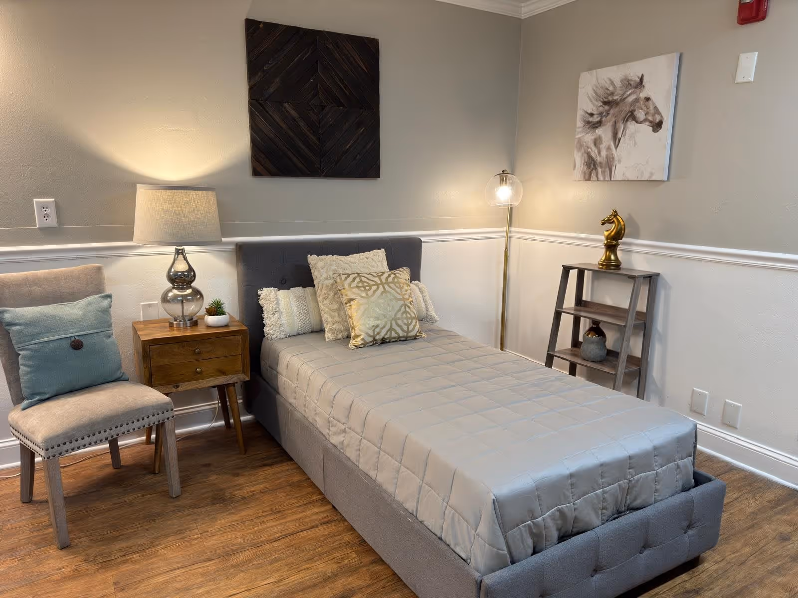 A neatly made single bed with a gray upholstered headboard and gray quilted bedspread, adorned with decorative pillows. Next to the bed is a wooden nightstand with a glass lamp and a small plant. A beige cushioned chair with a blue pillow is placed beside the nightstand. On the right side, there is a small wooden shelf with decorative items including a golden horse head sculpture, and above it hangs a painting of a horse. The room has wooden flooring and light gray walls with white trim.