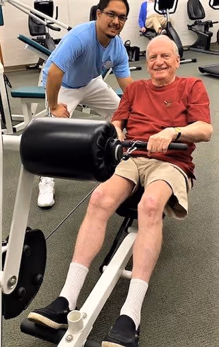 An elderly man in a red shirt and beige shorts is using a leg exercise machine in a fitness room. A younger man in a light blue shirt and white pants is standing beside him, smiling and providing support. Exercise equipment is visible in the background.