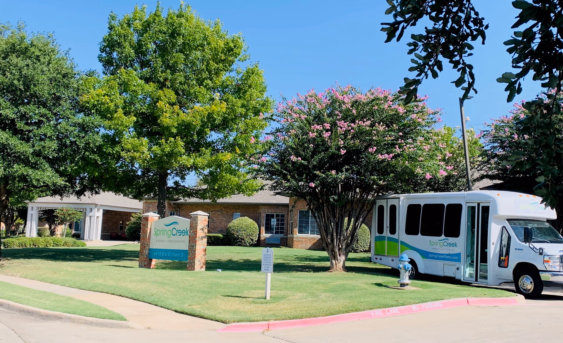Exterior front of the Spring Creek assisted living facility showing the entrance sign, shuttle van, brick building, and landscaped lawn with trees.