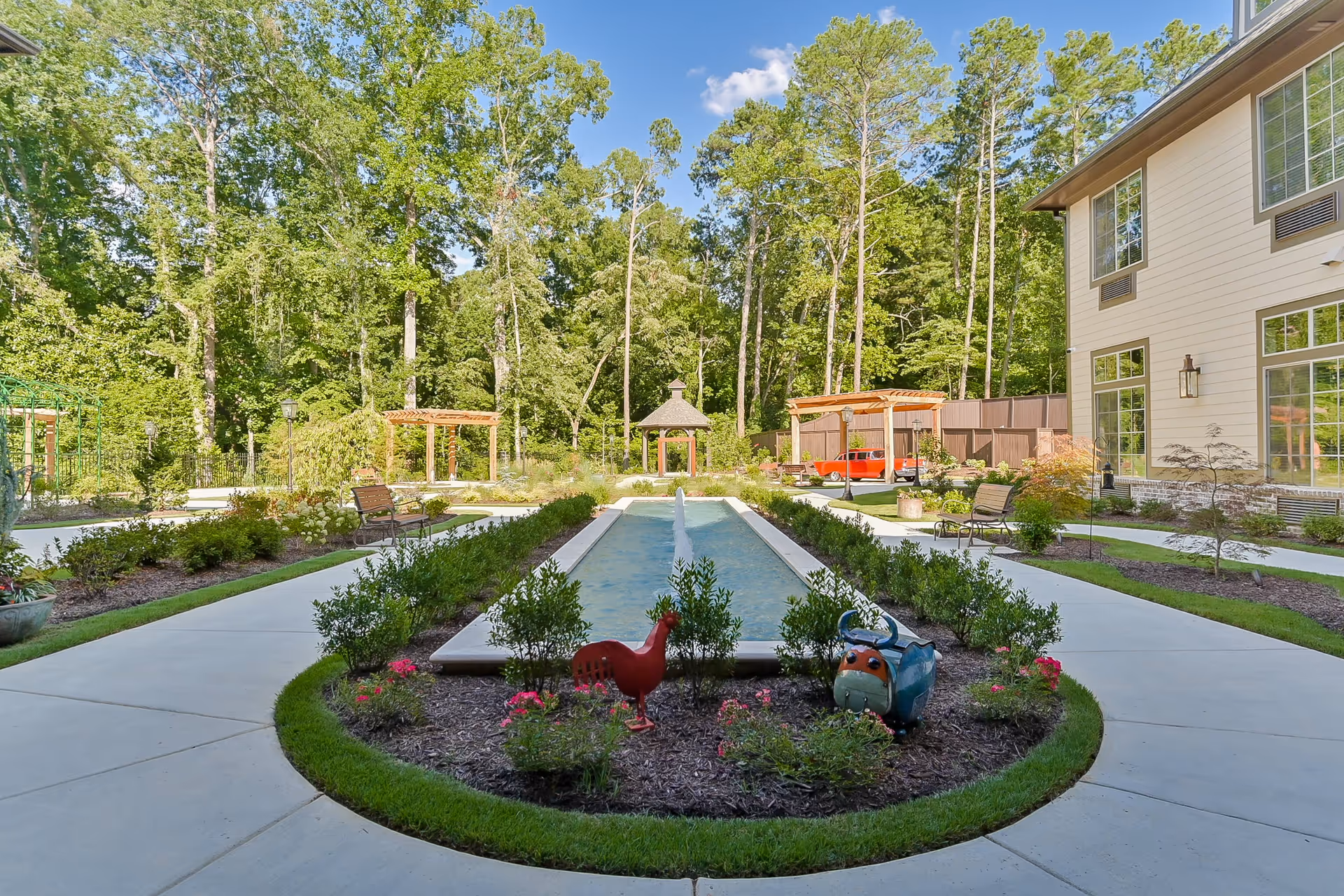 A landscaped outdoor garden area at Orchard at Brookhaven featuring a central rectangular water fountain with a small jet of water, surrounded by neatly trimmed bushes and flower beds. There are two decorative animal sculptures, one red and one blue, near the fountain. The garden has paved walkways, benches, wooden pergolas, and a gazebo in the background. Tall trees and a building with large windows are visible on the right side under a clear blue sky.