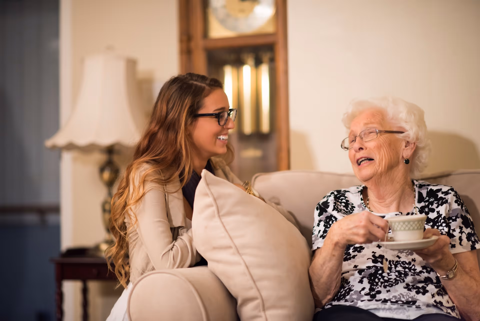 A young woman with long hair and glasses smiling and talking with an elderly woman who is holding a teacup and saucer while sitting on a beige couch in a cozy living room setting with a lamp and a grandfather clock in the background.