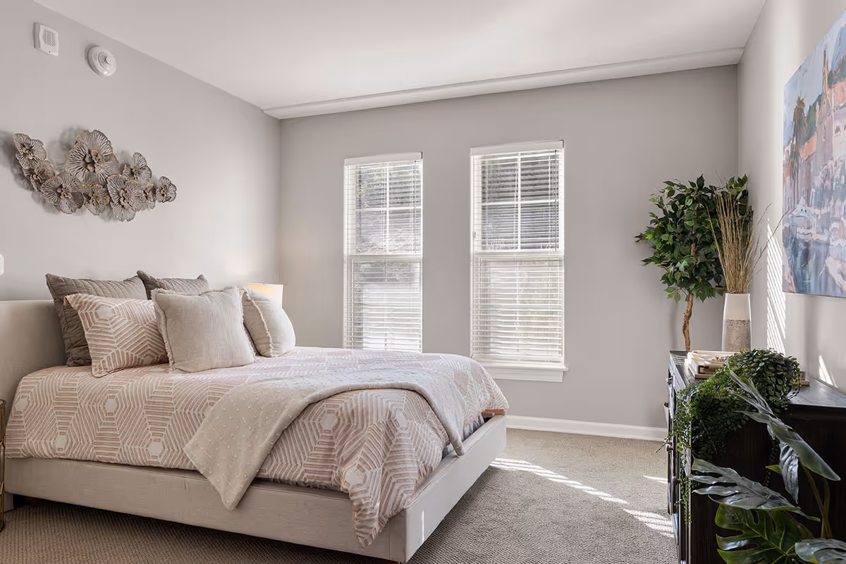 A bright bedroom with a neatly made bed featuring patterned bedding and multiple pillows. The room has two large windows with blinds, a decorative metal wall art above the bed, a dark wooden dresser with plants and books on top, and a colorful painting on the wall. The walls are painted light gray and the floor is carpeted.