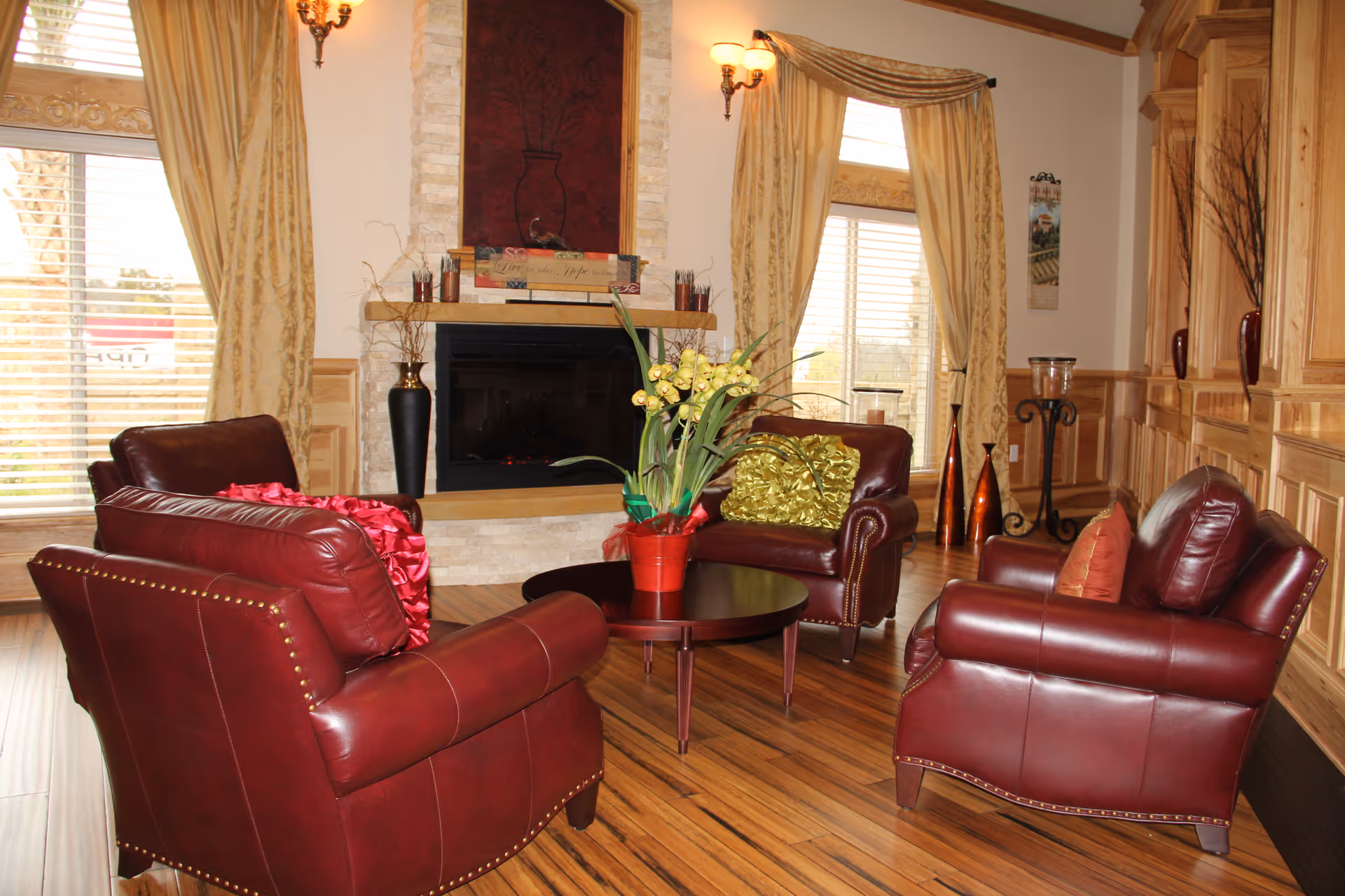 Cozy living room with four red leather armchairs arranged around a round coffee table in front of a fireplace, wood-paneled walls, and large draped windows.