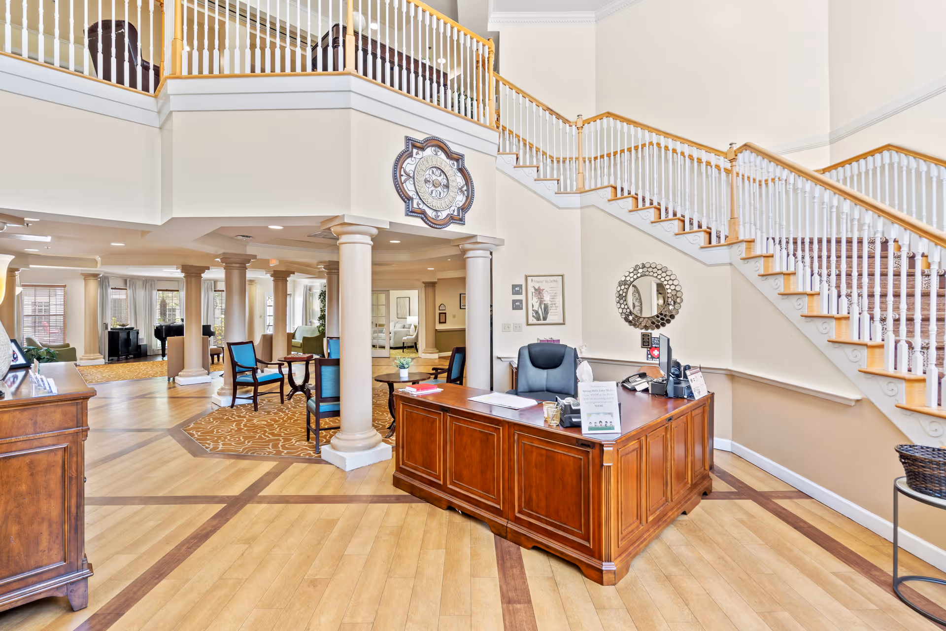 Bright and spacious senior living facility lobby with a wooden reception desk, a comfortable office chair, decorative columns, a staircase with white railings, and seating areas with blue chairs and small tables. The area is well-lit with natural light coming through large windows.