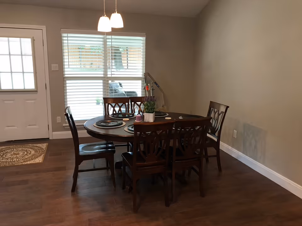 Dining area with a round wooden table set for six, matching chairs, pendant lights, a window with blinds, and a nearby entry door.