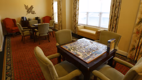 Cozy common room with upholstered chairs around tables, a partially completed jigsaw puzzle on one table, and a window seat.