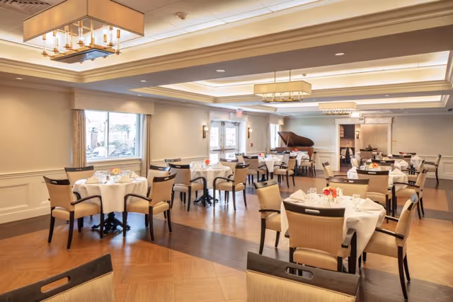 Bright, elegant dining room with round tables set for service, upholstered chairs, hardwood floors, and a grand piano.