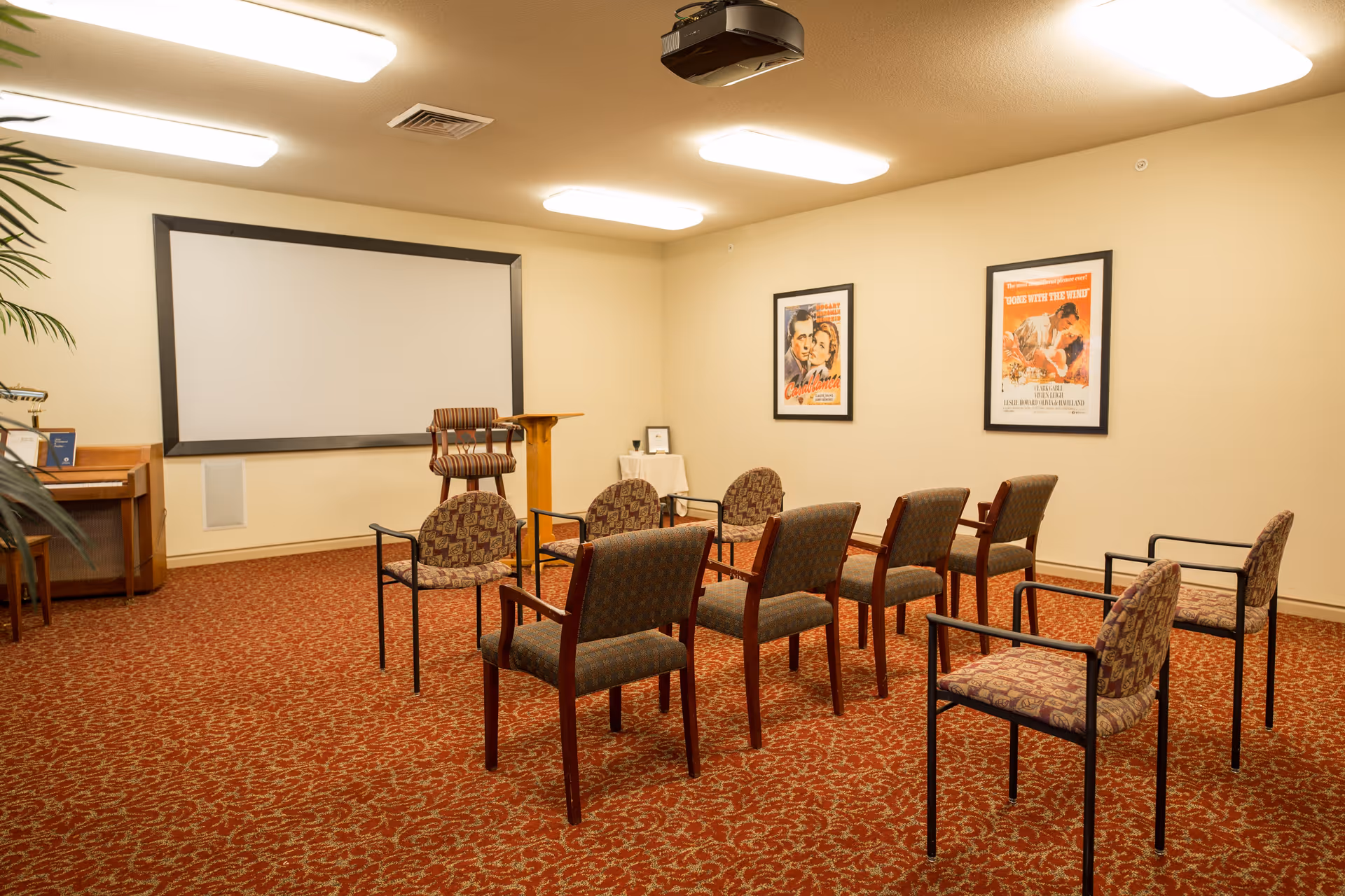 A small theater or presentation room with a large blank projection screen on the wall, several patterned chairs arranged in rows facing the screen, a wooden podium, and framed vintage movie posters on the wall. The room has beige walls and a red patterned carpet.