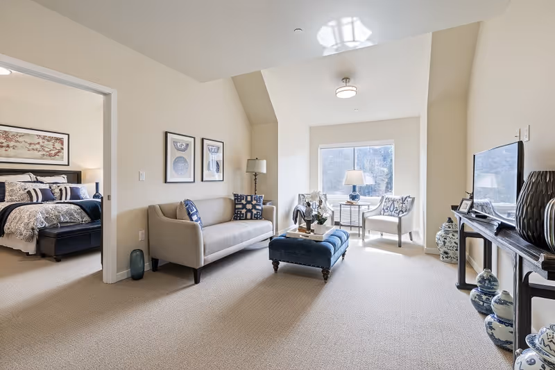A bright living room area with a beige sofa adorned with patterned pillows, a blue upholstered ottoman in the center, and two white armchairs near a large window letting in natural light. A floor lamp stands beside the sofa, and a TV is placed on a console table decorated with blue and white ceramic vases. Through an open doorway, a bedroom with a neatly made bed and framed artwork above the headboard is visible.