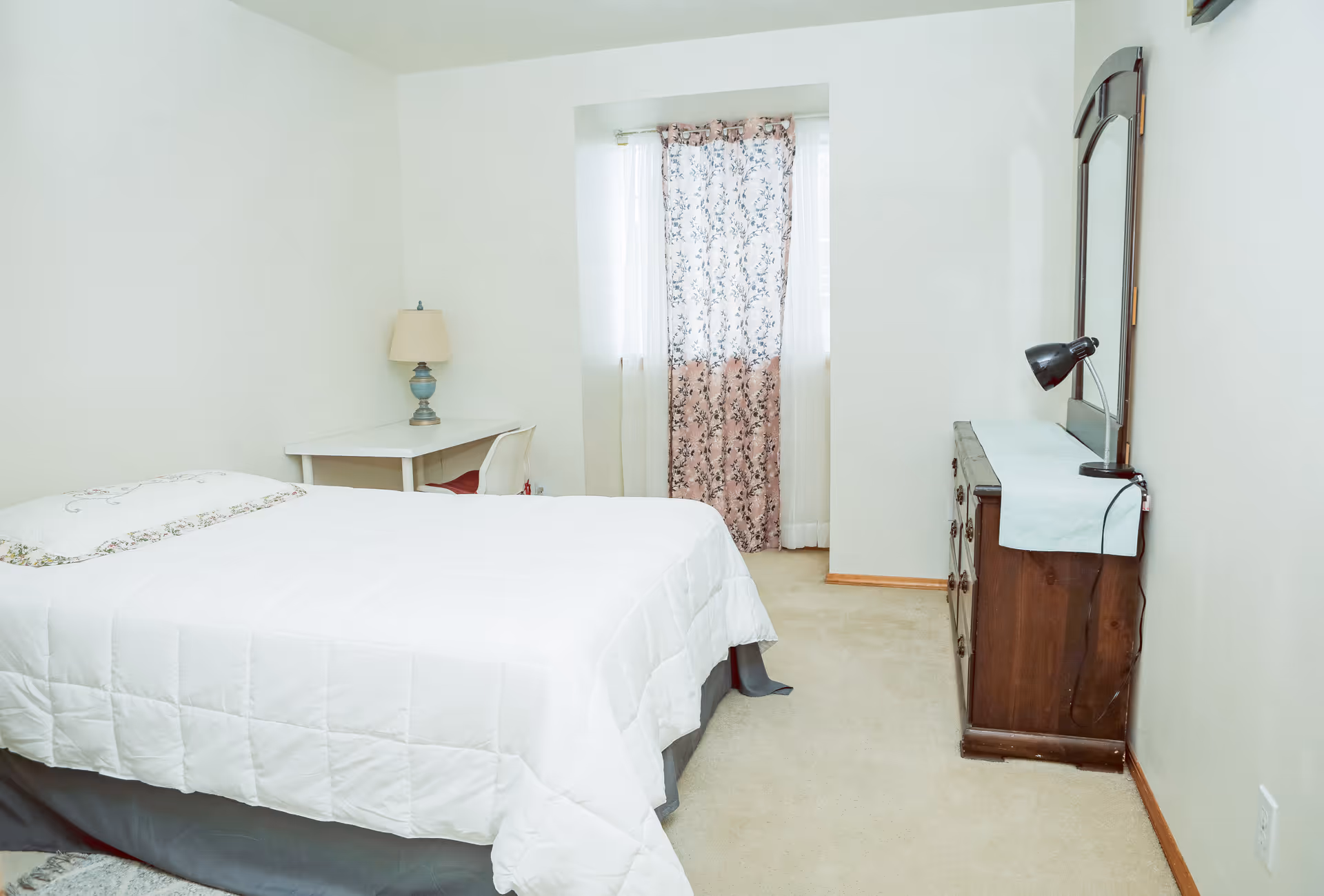 A bedroom with a neatly made bed covered in a white quilt. There is a small white desk with a lamp and chair in the corner near a window with floral curtains. On the right side, there is a wooden dresser with a mirror and a black desk lamp on top. The room has light-colored walls and carpeted floor.