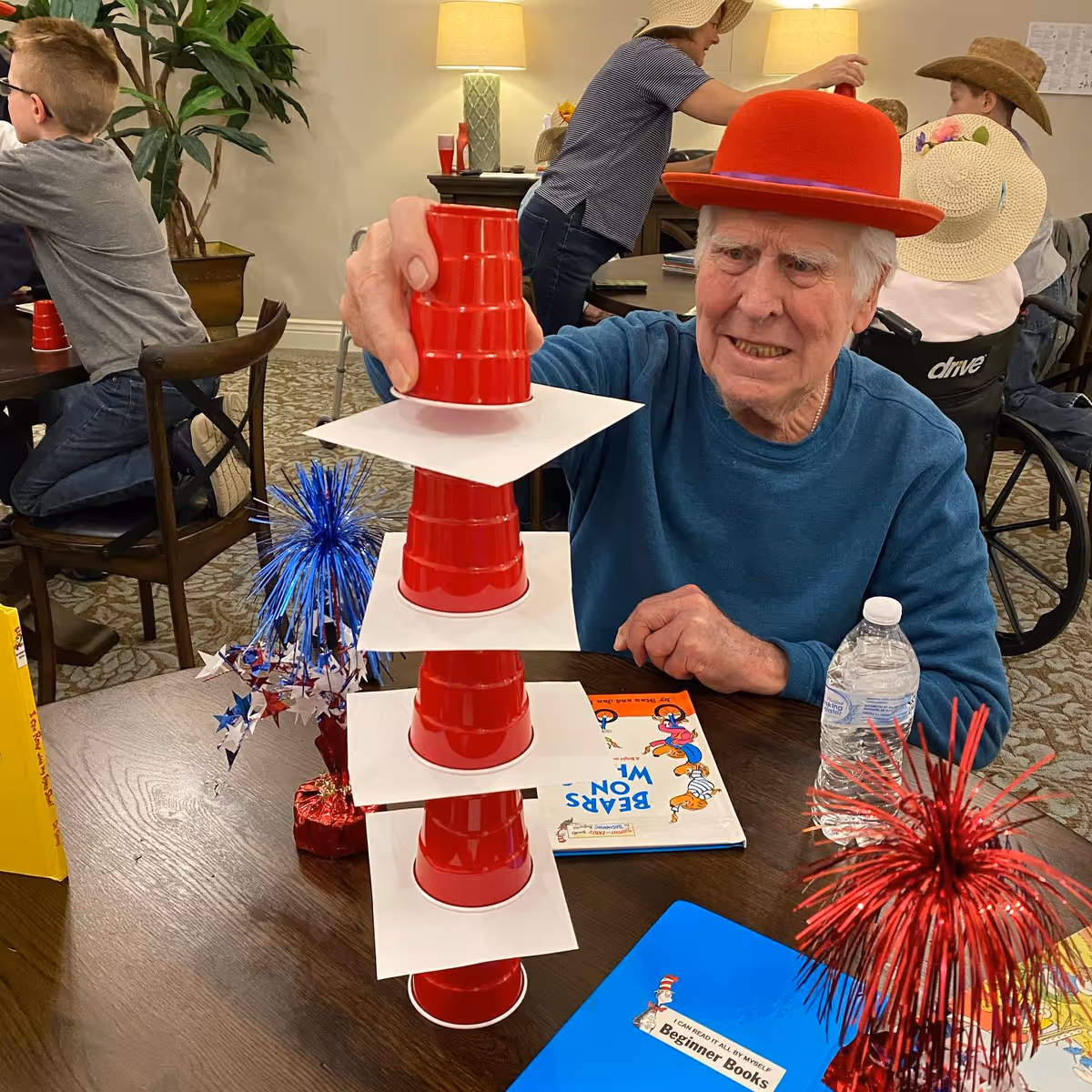 An elderly man wearing a red hat is carefully stacking red plastic cups separated by white square cards on a table. The table also has a water bottle, a book titled 'Bears on Wheels,' and red and blue decorative centerpieces. In the background, other people, including children wearing hats, are engaged in activities in a well-lit room with lamps and plants.