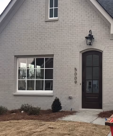 Exterior view of a light gray brick building with a large window and a dark wooden door. The building has the number 5009 displayed vertically next to the door, a black outdoor lantern above the door, and some small shrubs planted in front of the building.