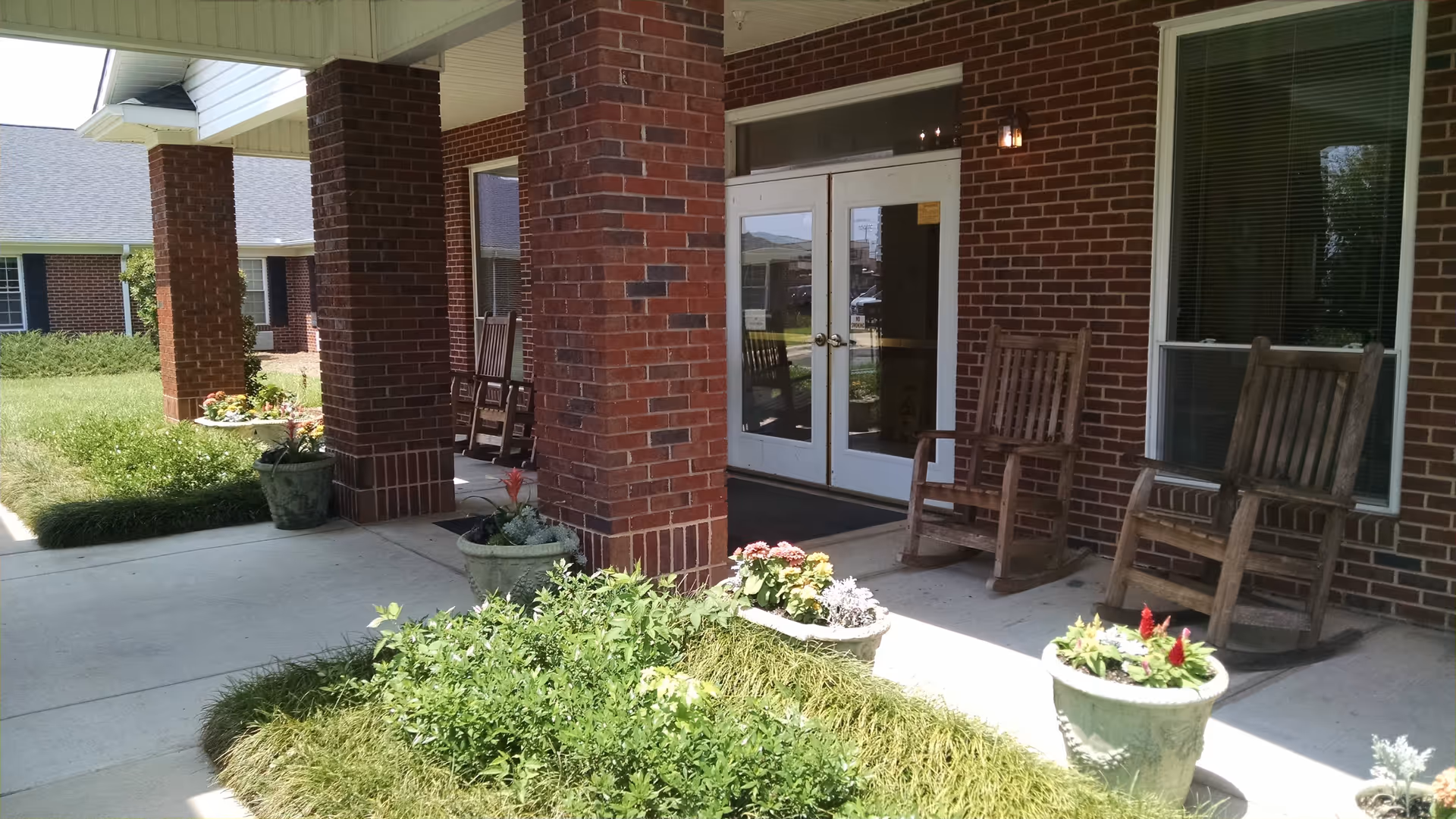 Covered entrance area of a brick building with two wooden rocking chairs on the porch, potted plants with flowers, and a glass double door entrance. Green grass and shrubs surround the walkway leading to the entrance.