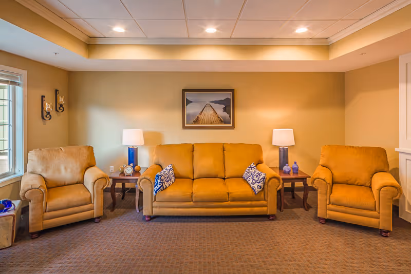 A cozy living room with a mustard yellow sofa and two matching armchairs. The sofa has two blue and white patterned throw pillows. There are two wooden side tables with blue lamps on either side of the sofa. A framed picture of a wooden pier extending into a calm body of water hangs on the beige wall behind the sofa. The room has a carpeted floor and recessed ceiling lights.