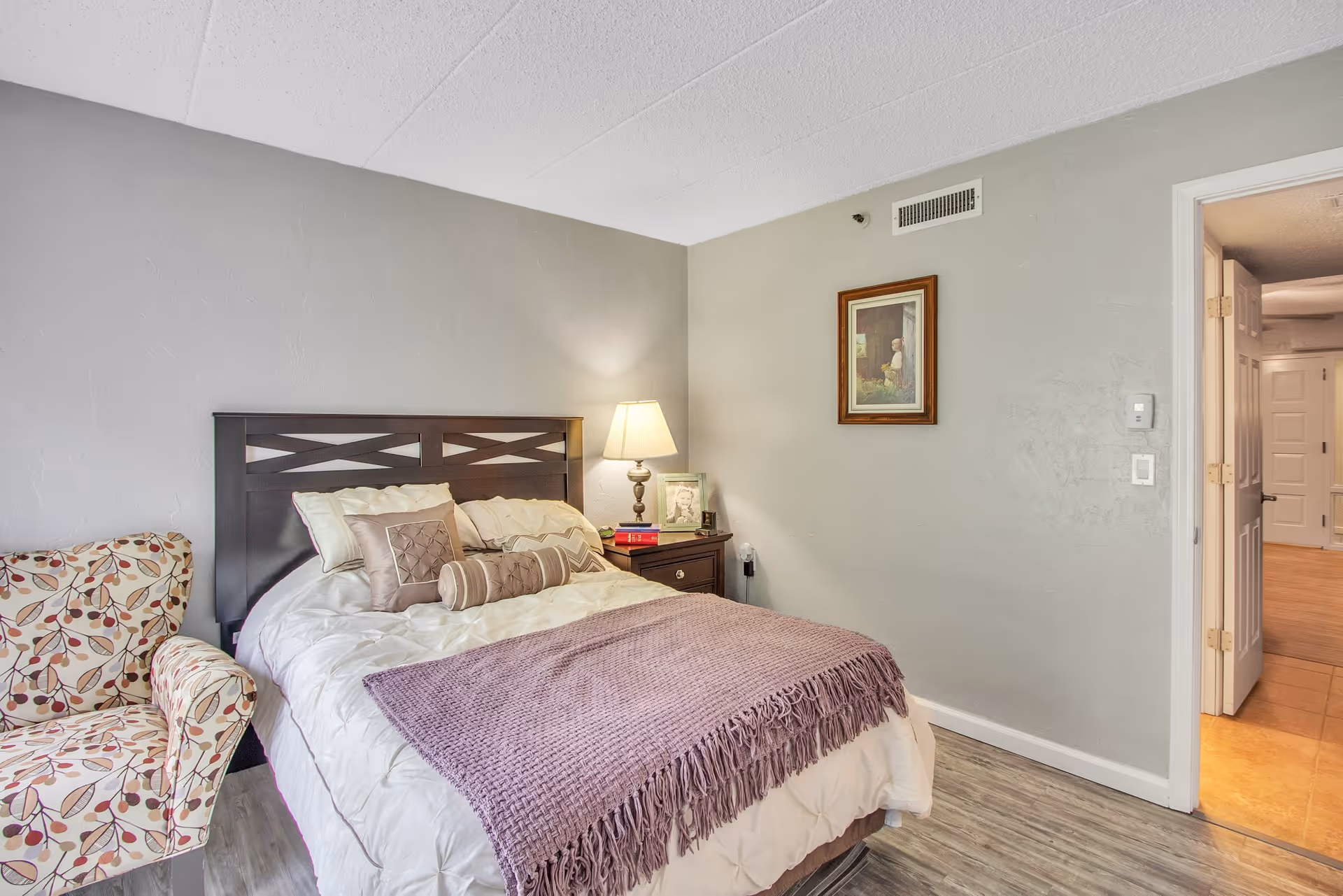 A cozy bedroom with a bed featuring a dark wooden headboard, white bedding, and multiple decorative pillows. A purple throw blanket is draped across the foot of the bed. To the left of the bed is a patterned armchair with a leaf design. A nightstand with a lamp, books, and framed photos is positioned next to the bed. A framed picture hangs on the wall above the nightstand. The room has light gray walls and wood-look flooring. An open door reveals a hallway with tiled flooring.