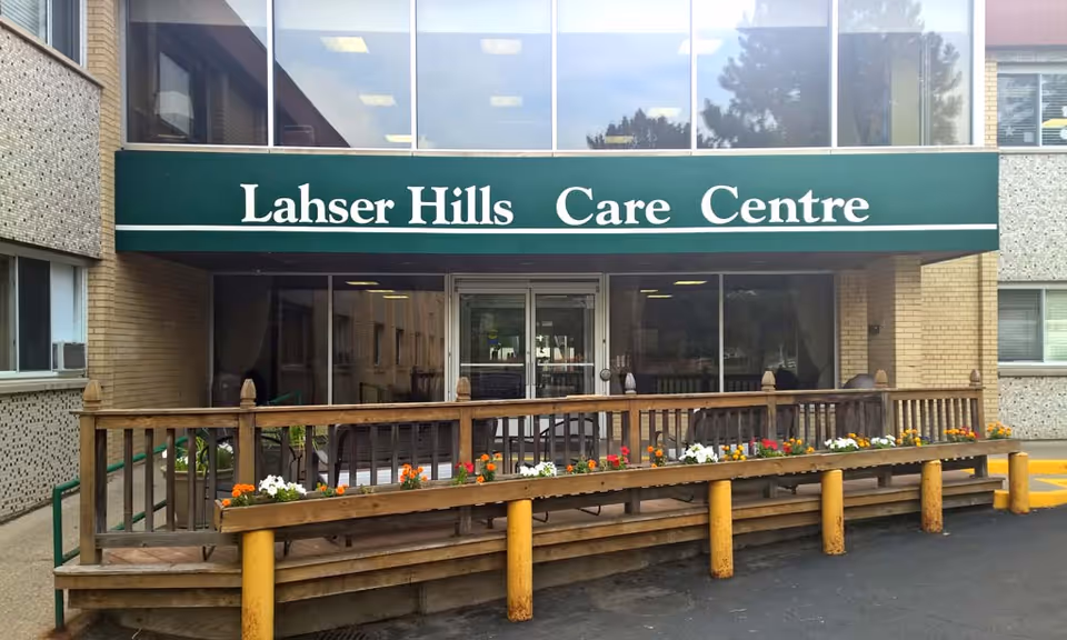 Entrance of Lahser Hills Care Centre with a green awning displaying the facility name. There is a wooden ramp with flower boxes containing colorful flowers leading to the glass double doors of the building.