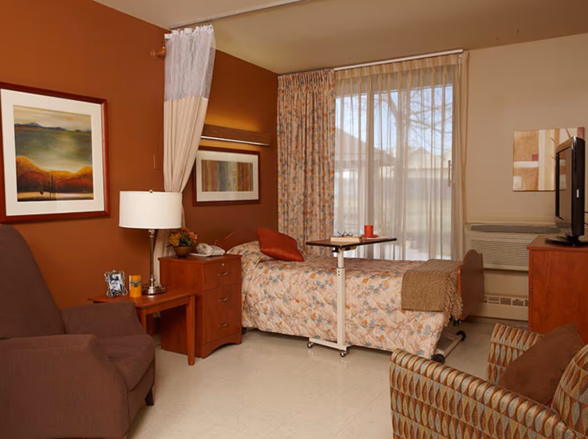 A cozy senior living bedroom with a single bed covered in a floral quilt, a brown armchair, a patterned armchair, a wooden nightstand with a lamp and telephone, framed artwork on the walls, and a large window with sheer and floral curtains letting in natural light.