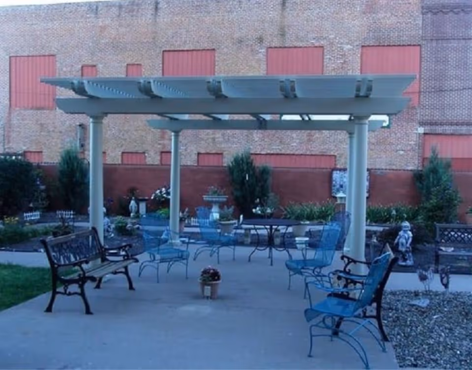 Outdoor seating area with metal benches and chairs arranged under a white pergola. The area is paved with concrete and surrounded by garden beds with plants and shrubs. A brick building with red boarded-up windows is visible in the background.