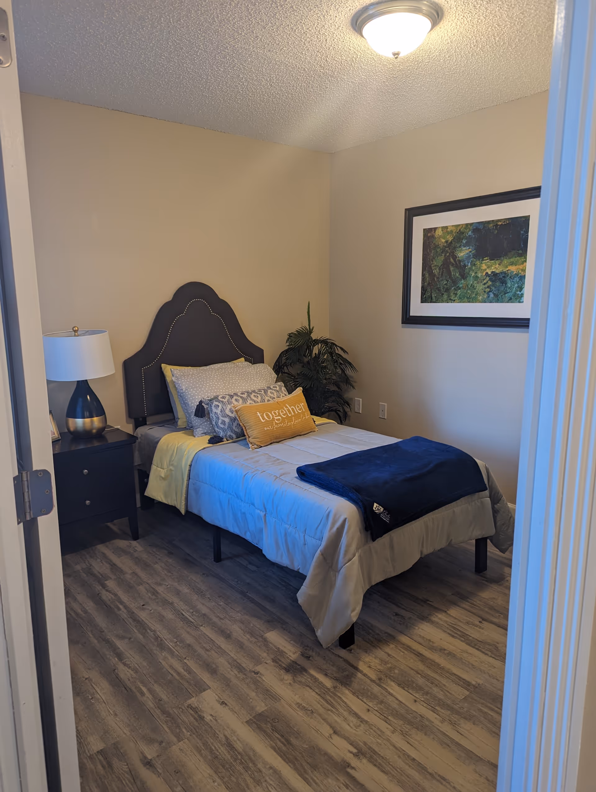 A small bedroom with a single bed featuring a dark upholstered headboard. The bed is made with light-colored bedding, multiple pillows including one with the word 'together' on it, and a dark blue blanket folded at the foot. There is a black nightstand with a lamp on it to the left of the bed, a green potted plant in the corner, and a framed landscape painting on the wall to the right. The floor is wood-style laminate, and the walls are painted beige.