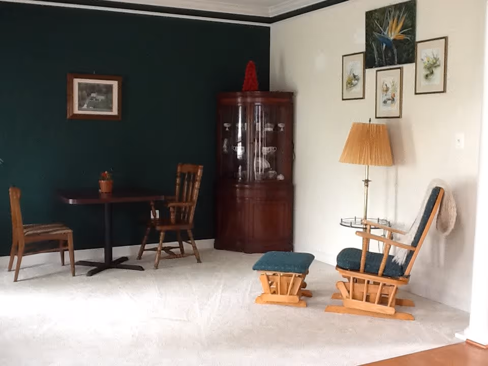 A cozy corner of a living room with a small wooden table and two chairs on the left, a dark wooden corner cabinet with glass doors displaying dishes, and a wooden rocking chair with a matching footrest and a floor lamp on the right. The walls are painted dark green and white, decorated with framed pictures and a painting of a bird of paradise flower.
