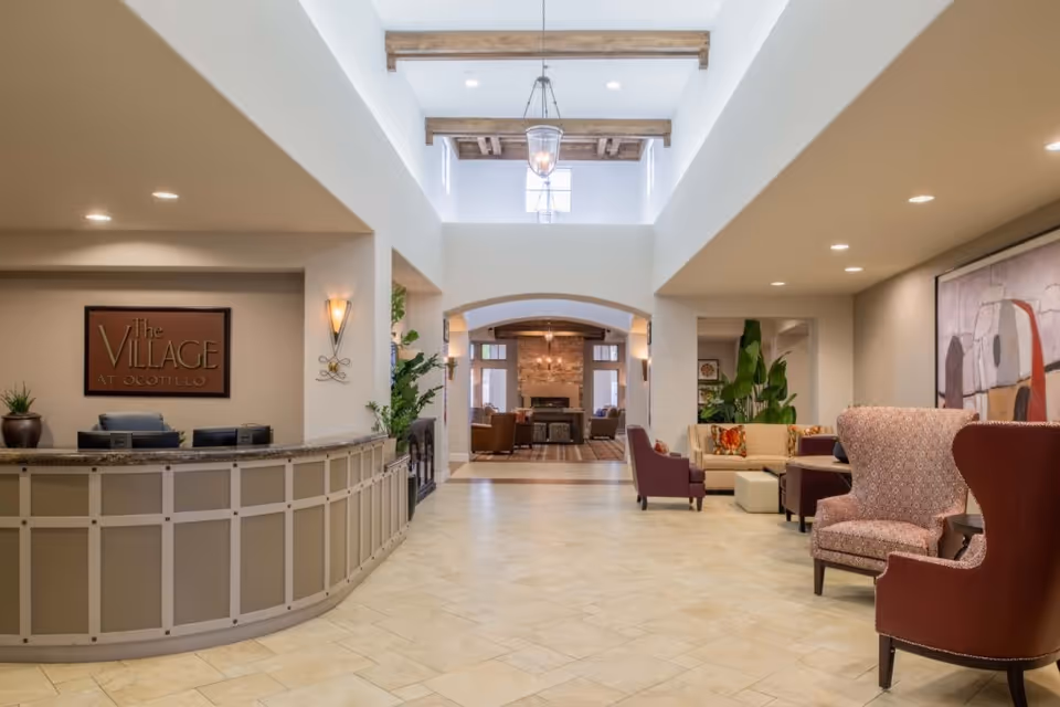 Reception lobby with a curved check-in desk, seating areas, high ceiling skylight, and a wall sign reading "The Village at Ocotillo".