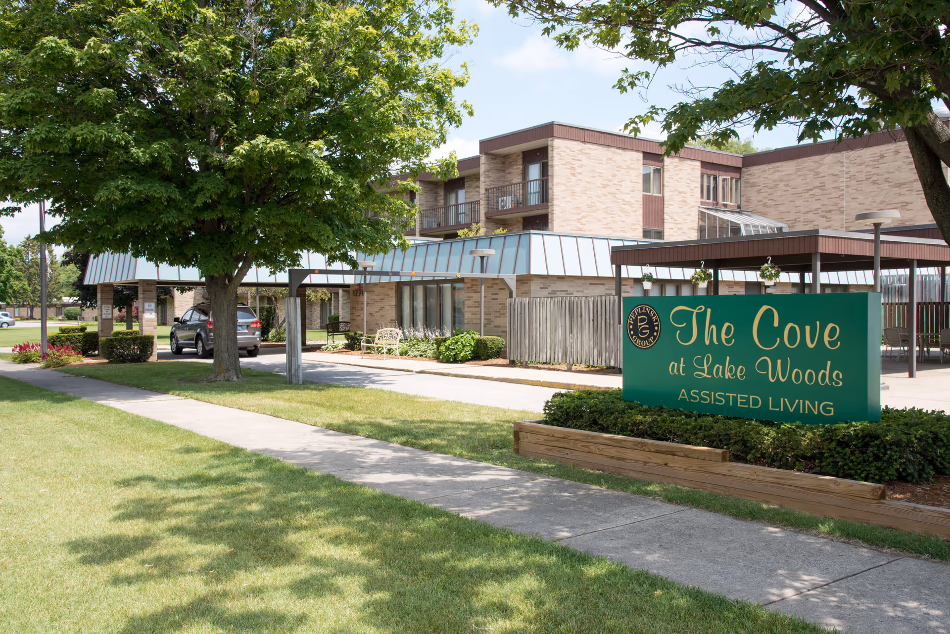 Exterior front entrance of The Cove at Lake Woods assisted living center showing the building, driveway, landscaping, and a green sign.