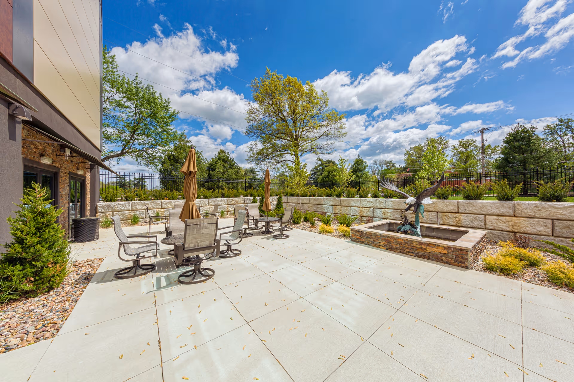 Outdoor patio area at St. Anthony’s Senior Living with several round tables and chairs, closed umbrellas, a stone planter with a large eagle sculpture, surrounded by greenery and trees under a partly cloudy blue sky.