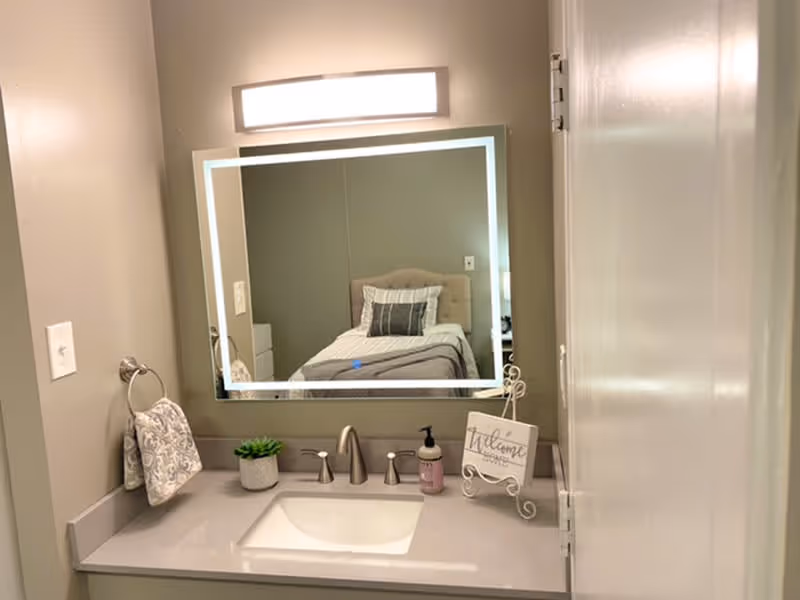 A bathroom sink area with a modern faucet, a small potted plant, a soap dispenser, and a decorative sign that says 'Welcome'. Above the sink is a large rectangular mirror with a light fixture above it. The mirror reflects a bed with pillows and a headboard in an adjoining room.