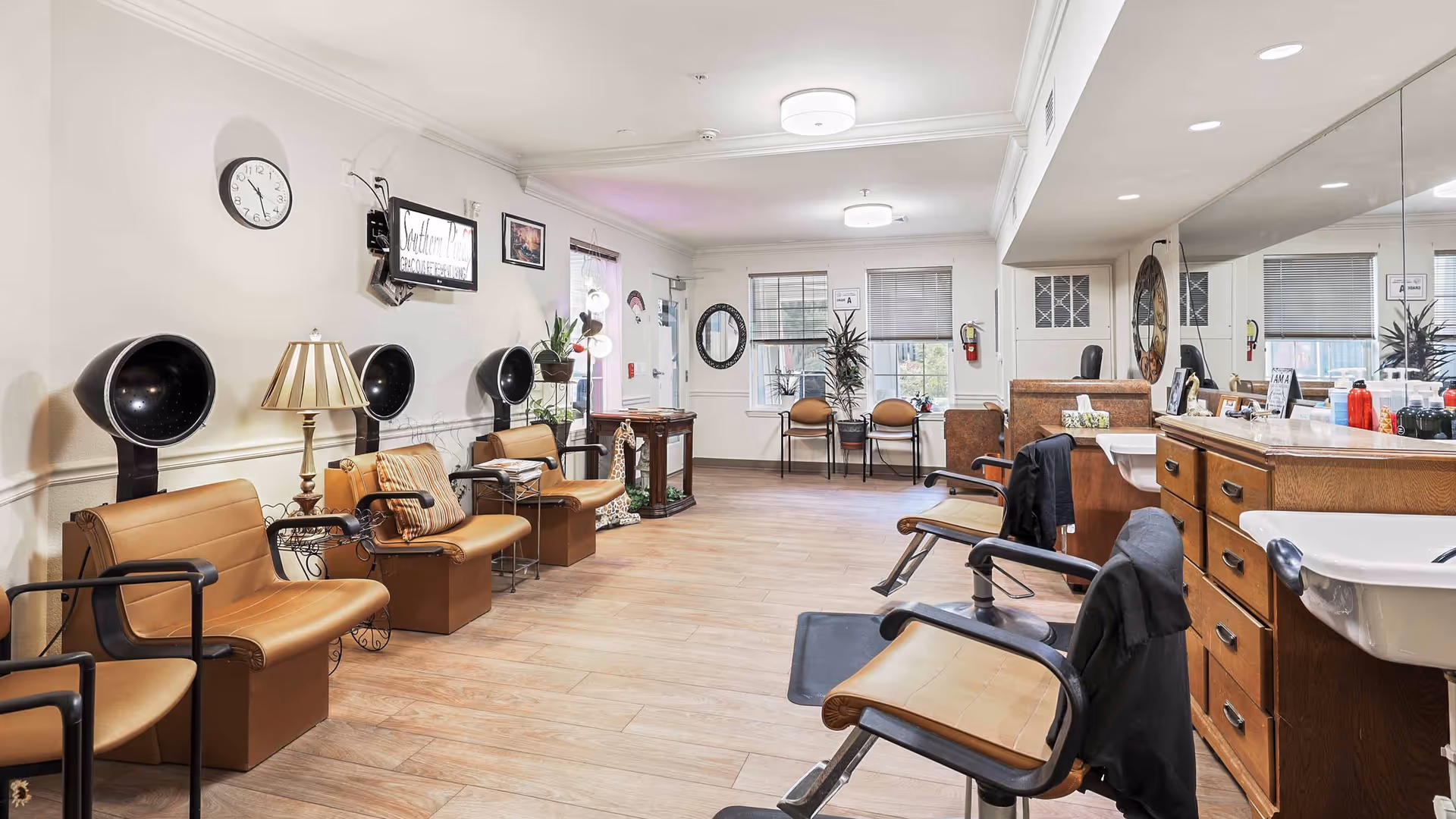 Interior view of a hair salon area in a senior living facility with several brown salon chairs, hair dryers mounted on the wall, a large mirror, sinks, and various hair care products on the counter. The room has light-colored walls, wood flooring, and windows letting in natural light.