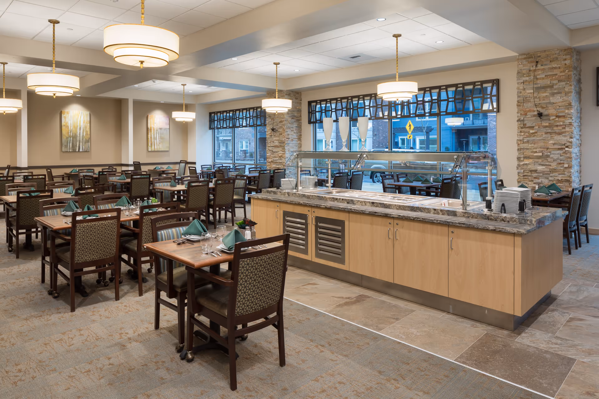 A spacious dining room in a senior living facility with multiple wooden tables and chairs neatly arranged. Each table is set with green folded napkins, glasses, and silverware. In the center, there is a buffet counter with a marble countertop and a glass sneeze guard. Large windows with decorative valances allow natural light to enter, and modern pendant lights hang from the ceiling. The walls feature stone accents and framed artwork.