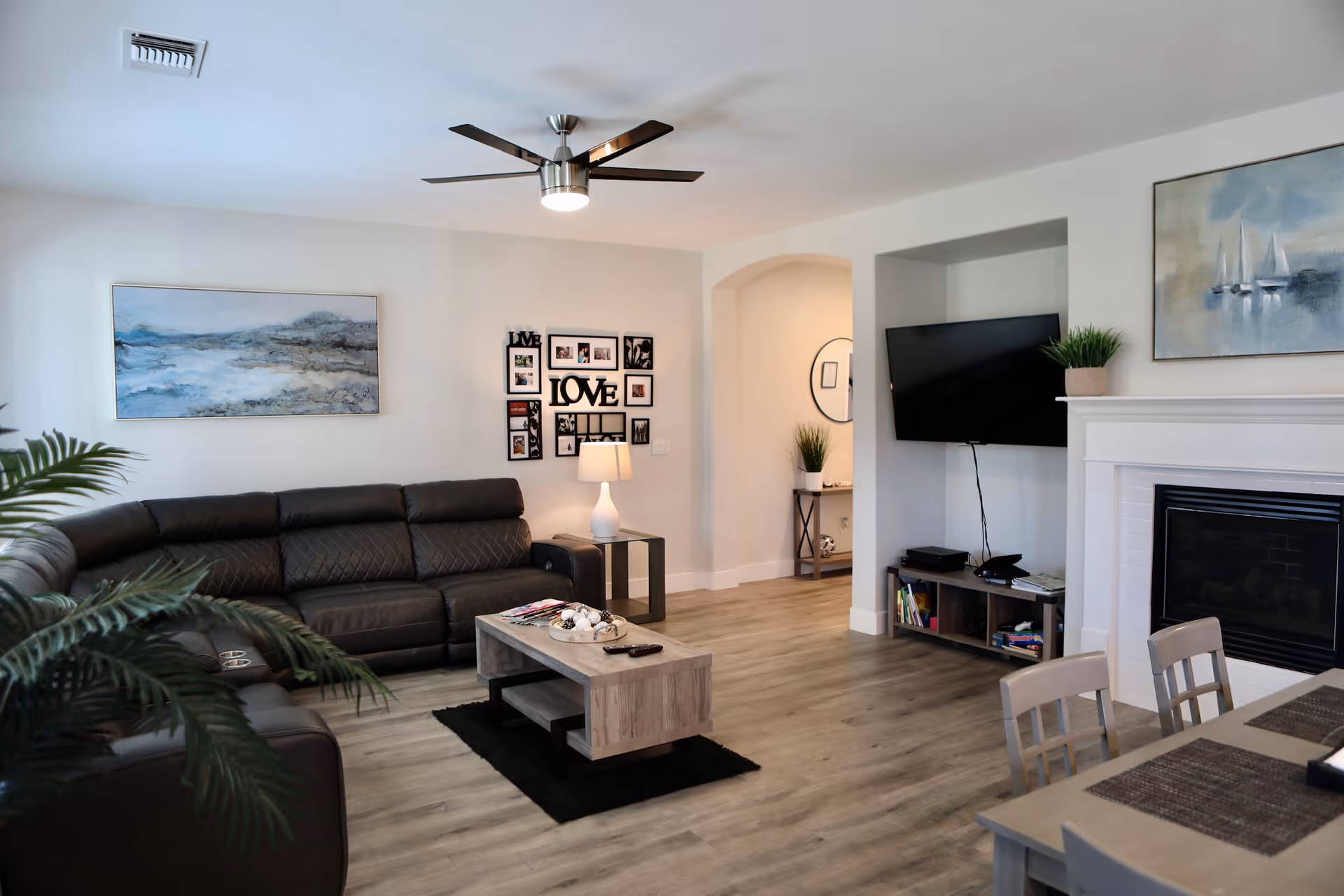A modern living room with a dark brown leather sectional sofa, a wooden coffee table on a black rug, and a flat-screen TV mounted on the wall above a small entertainment unit. The room features light wood flooring, a white fireplace with a painting above it, a ceiling fan with lights, and a dining table with chairs in the foreground. There are decorative plants and framed pictures on the walls.