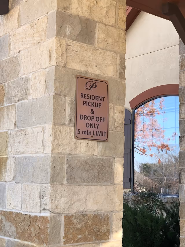 A stone pillar with a sign that reads 'Resident Pickup & Drop Off Only 5 min Limit' at the entrance of a building with a window reflecting trees and sky.