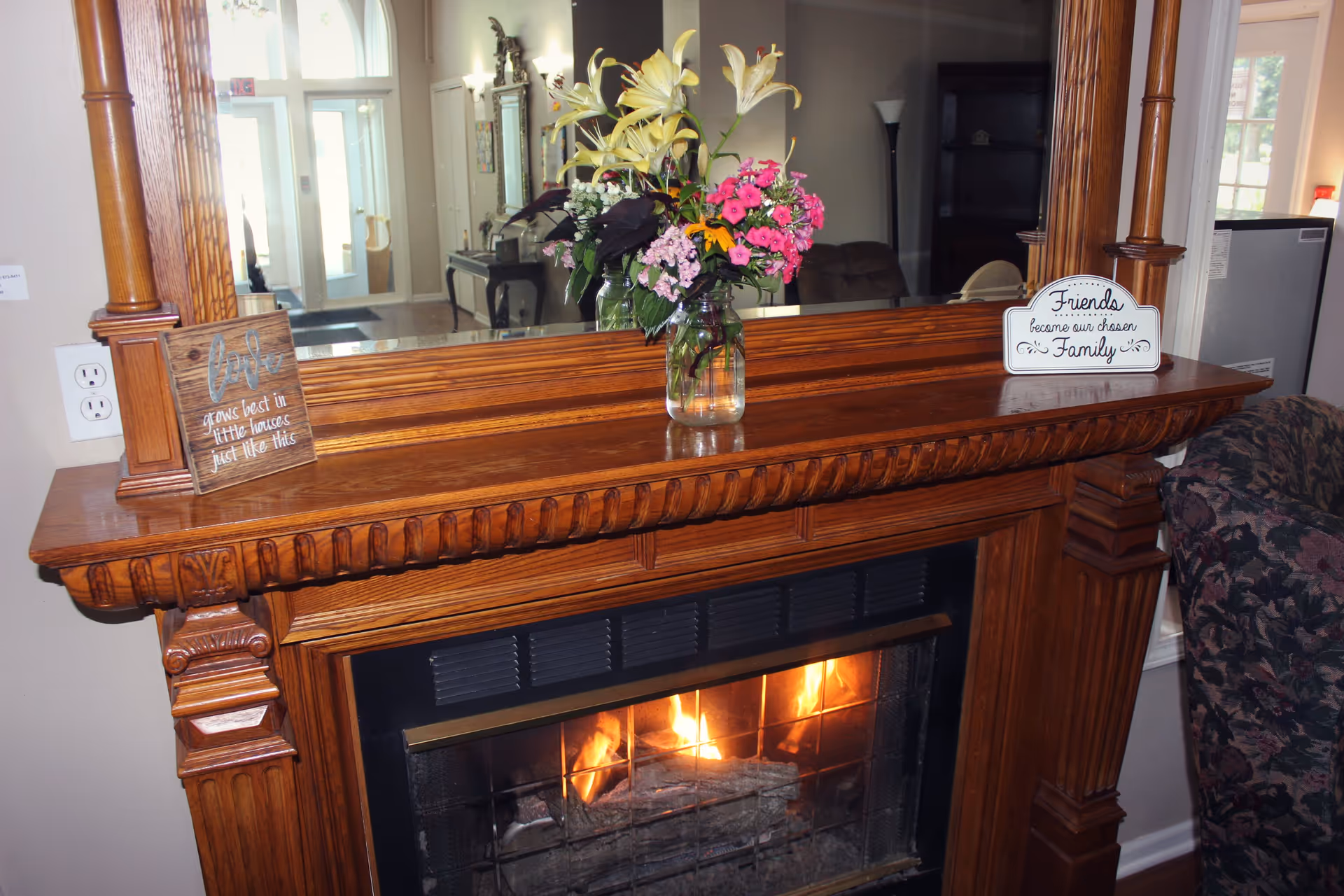 A cozy wooden fireplace mantel with a burning fire inside. On top of the mantel, there is a glass jar with a bouquet of colorful flowers, a small wooden sign that reads 'love grows best in little houses just like this,' and another sign that says 'Friends become our chosen Family.' A large mirror is mounted above the mantel reflecting part of the room, including a doorway and furniture.