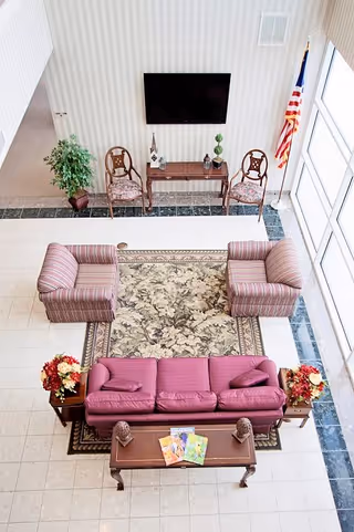 A bright and spacious seating area with a purple sofa, two striped armchairs, a floral patterned rug, and two wooden side tables with flower arrangements. Behind the seating area are two wooden chairs with cushions, a small table with decorative items, a flat-screen TV mounted on the wall, and an American flag near large windows letting in natural light.