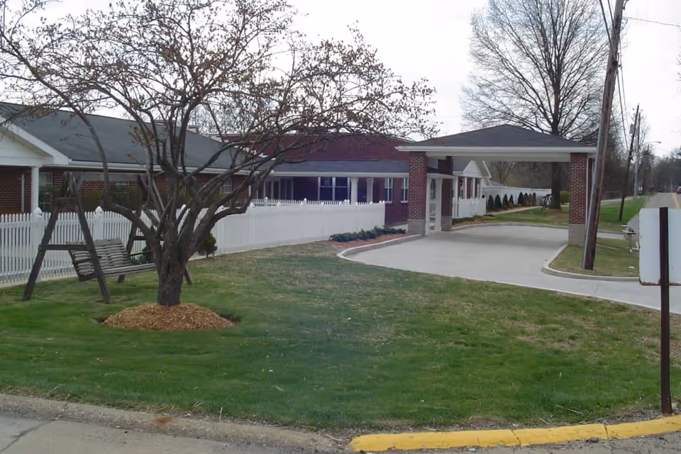 Front exterior of a single-story brick health center with a covered drop-off entrance, white picket fence, lawn, and a tree with a wooden swing.