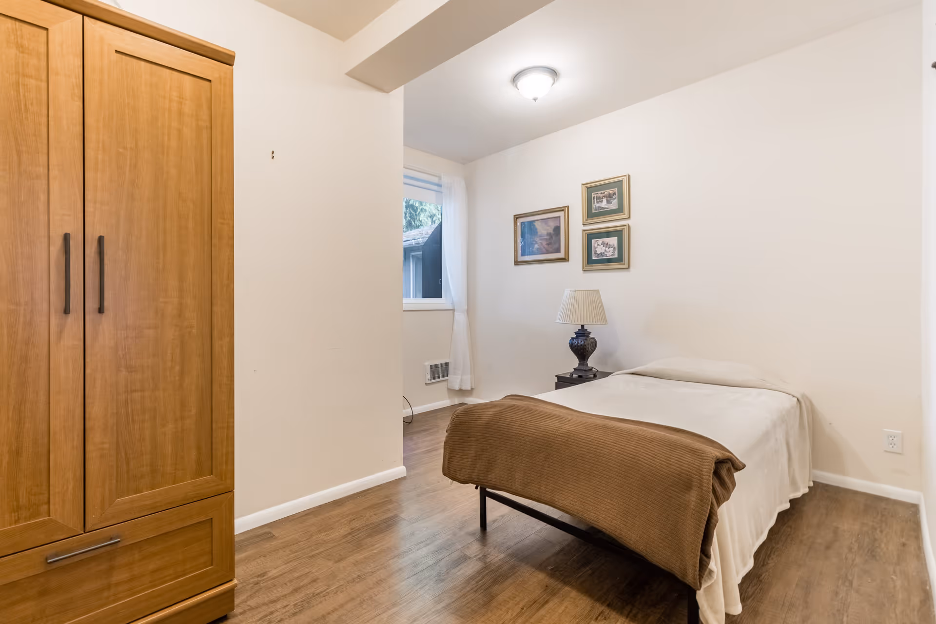 A small bedroom with a single bed covered in white sheets and a brown blanket at the foot. Next to the bed is a dark nightstand with a decorative lamp. Three framed pictures hang on the white wall above the nightstand. A wooden wardrobe is on the left side of the image, and a window with sheer white curtains is visible in the background. The floor is wooden, and the room has a simple, clean design.