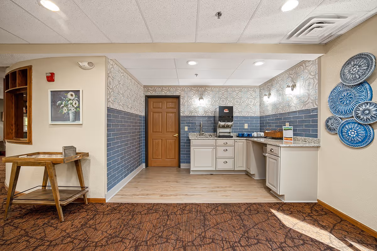 Small kitchenette in a senior living common area with a sink, coffee machine, cabinets, and decorative wall plates.