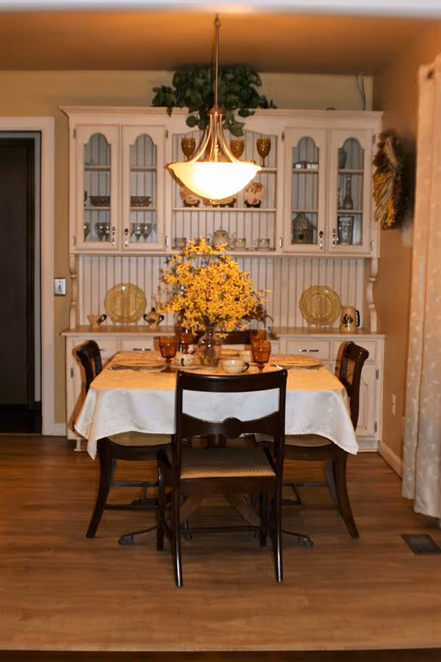 A cozy dining room with a wooden table covered by a white tablecloth, set with amber-colored glasses, plates, and a centerpiece of yellow flowers in a vase. Four wooden chairs surround the table. Behind the table is a white hutch with glass doors displaying dishes and decorative items. A hanging light fixture illuminates the table, and beige curtains are visible on the right side.