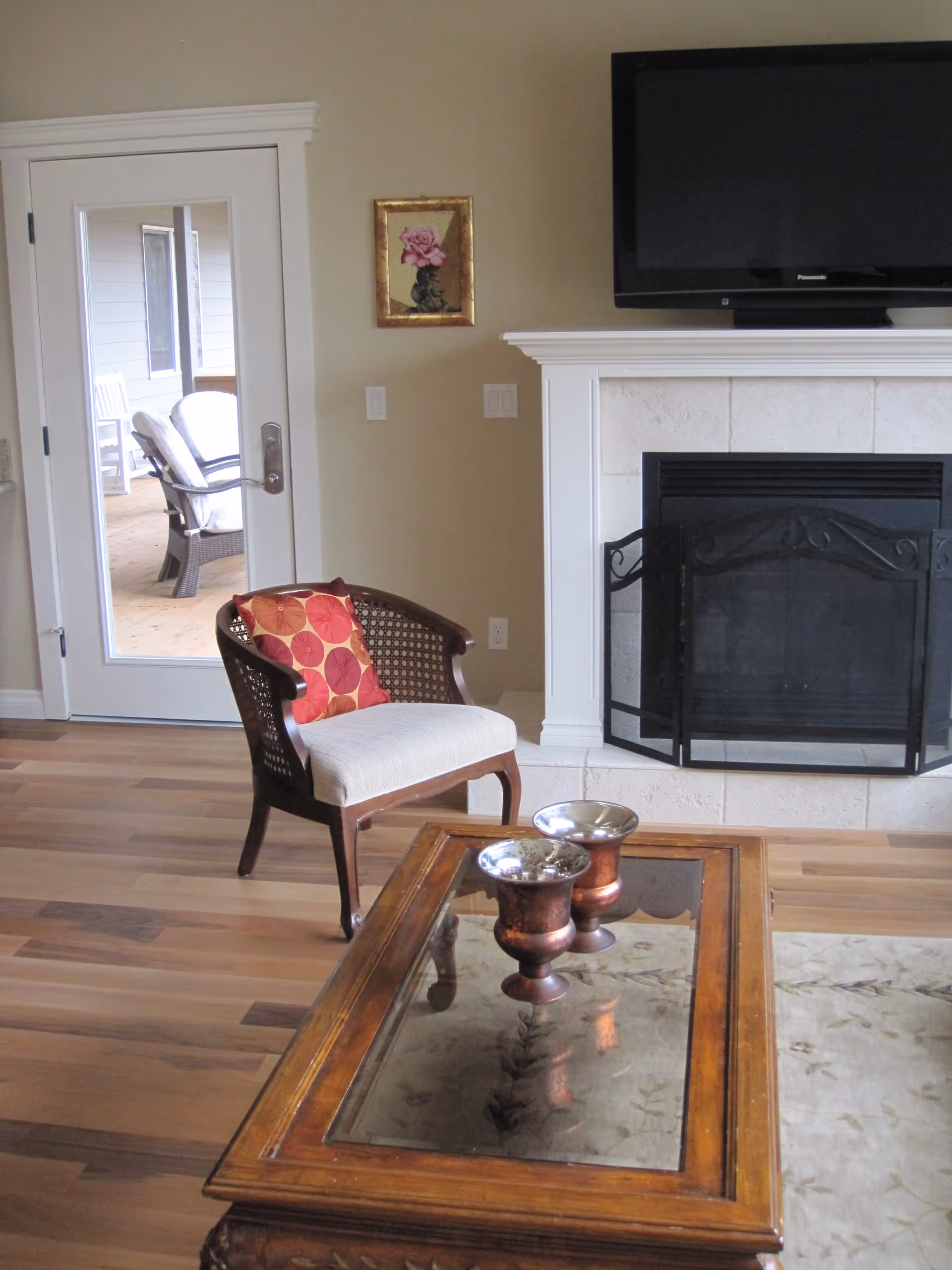 A cozy living room with a wooden chair featuring a red and orange patterned cushion, a wooden coffee table with a glass top holding two decorative metallic vases, a fireplace with a black screen, and a wall-mounted flat-screen TV above the fireplace. A glass door leads to an outdoor patio with cushioned chairs visible.
