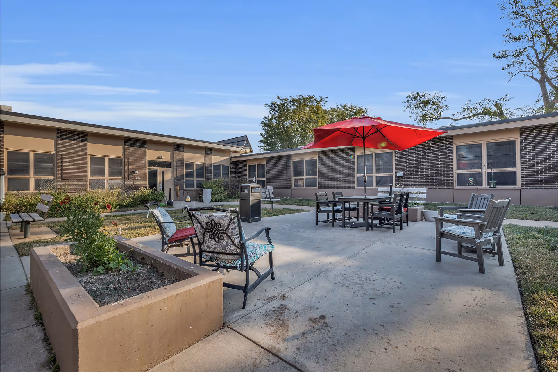 Outdoor courtyard area of Kenwood View Health and Rehabilitation Center featuring a concrete patio with several chairs and a table under a large red umbrella. Surrounding the patio are garden beds, benches, and a single-story brick building with multiple windows. Trees and a clear blue sky are visible in the background.