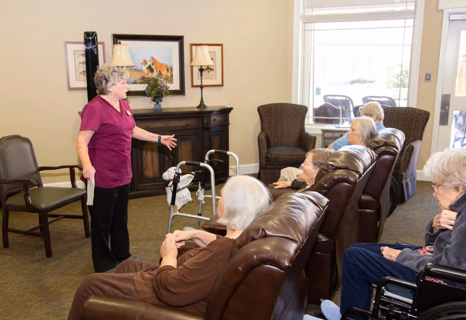 A group of elderly residents seated in a living room area with leather recliners and chairs, attentively listening to a staff member in a purple uniform who is standing and speaking to them. The room has a cabinet with lamps and framed pictures on the wall, and large windows letting in natural light.