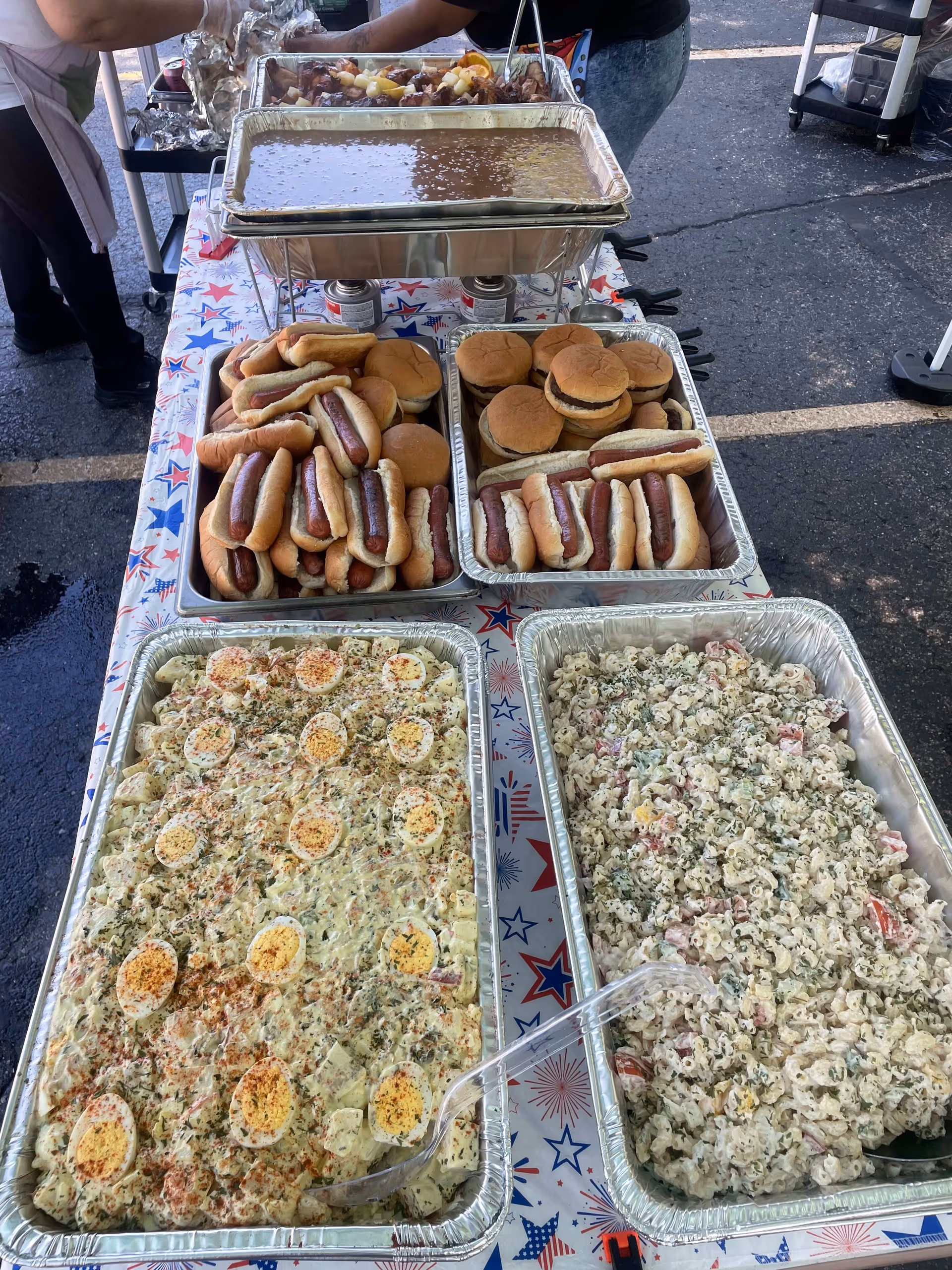 A buffet table outdoors with trays of food including hot dogs in buns, hamburgers, a tray of baked beans, potato salad topped with sliced boiled eggs and herbs, and a macaroni salad with vegetables. Two people are serving food in the background.